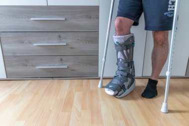 Achilles tendon yırtılması ameliyatından sonra Avrupalı bir adam evinde ay botu özel fizyoterapi ayakkabısıyla ve yaralı bacağa karşı evde tedavi için koltuk değnekleriyle ilk adımları PWB olarak atmayı öğreniyor.