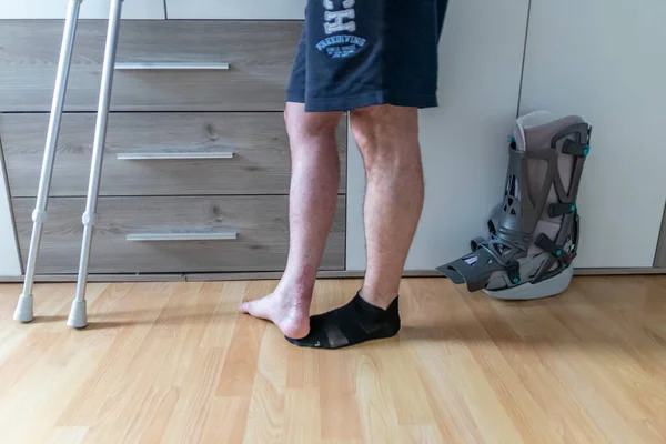 アキレス腱断裂手術後のヨーロッパの男は 最初のステップを歩くことを学ぶ痛み足に対して自宅で回復のための月のブーツ特別な理学療法の靴と松葉杖を家に帰っていますPwb — ストック写真