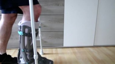 European man after Achilles tendon  rupture operation is back home with moon boot special physiotherapy shoe and crutches for recovery at home against the hurting leg learning to walk first steps PWB