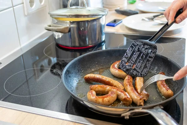 Woman cooking sausages in pan on kitchen cooking plate with fork and black spatula with delicious fatty bratwurst for dinner as unhealthy but spicy and roasted sausage meal for lunch and the children