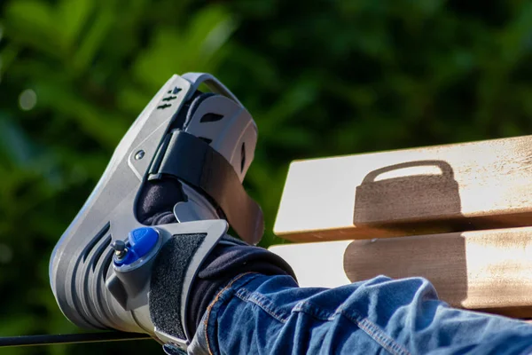 Boy with broken foot and orthopedic shoe or walker after bone fracture rests in public park on bench in green grass to recreate and rehabilitate his painful accident with medical equipment and plaster