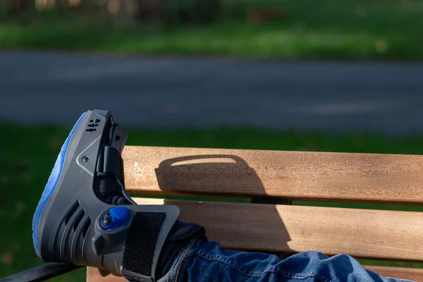 Boy with broken foot and orthopedic shoe or walker after bone fracture rests in public park on bench in green grass to recreate and rehabilitate his painful accident with medical equipment and plaster