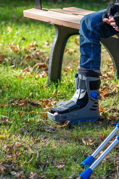 Boy with broken foot and orthopedic shoe or walker after bone fracture rests in public park on bench in green grass to recreate and rehabilitate his painful accident with medical equipment and plaster