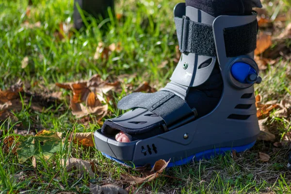 Boy with broken foot and orthopedic shoe or walker after bone fracture rests in public park on bench in green grass to recreate and rehabilitate his painful accident with medical equipment and plaster