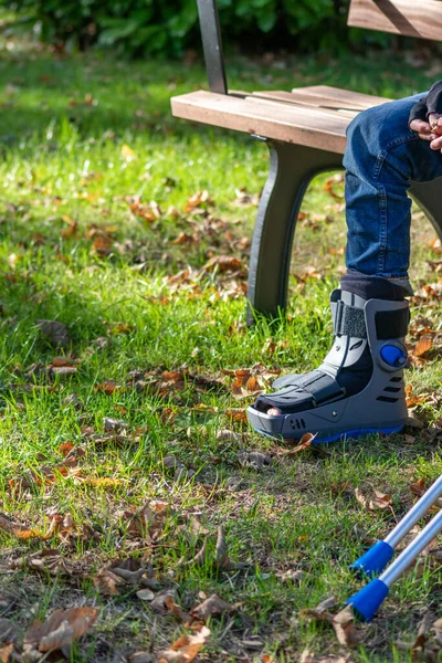 Boy with broken foot and orthopedic shoe or walker after bone fracture rests in public park on bench in green grass to recreate and rehabilitate his painful accident with medical equipment and plaster