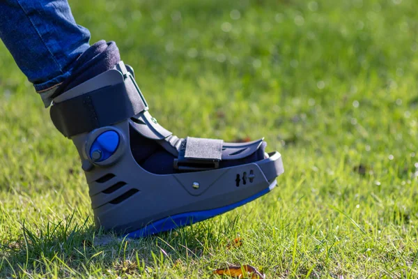 Boy with broken foot and orthopedic shoe or walker after bone fracture rests in public park on bench in green grass to recreate and rehabilitate his painful accident with medical equipment and plaster