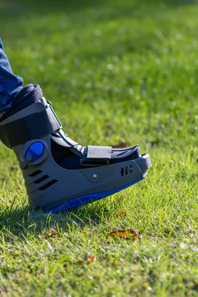 Boy with broken foot and orthopedic shoe or walker after bone fracture rests in public park on bench in green grass to recreate and rehabilitate his painful accident with medical equipment and plaster