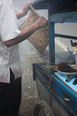 Satay satıcısı bir el arabası ile etrafta dolanıyor ve satayın düzgün ızgaralandığından emin oluyor. Doymak, bambu şiş ile tatlandırılmış et veya tavuktur..