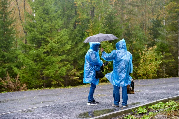 Ruskeala, Russia - 09.14.2021: Due persone in impermeabili blu sotto la pioggia in un parco autunnale a Ruskeala, Carelia. Un uomo tiene un ombrello sopra una donna mentre lei indossa i guanti. — Foto Stock