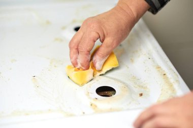 Bir kadın bir gaz sobasının yüzeyini sarı bir sünger, masa tuzu ve deterjan janıyla yıkıyor.