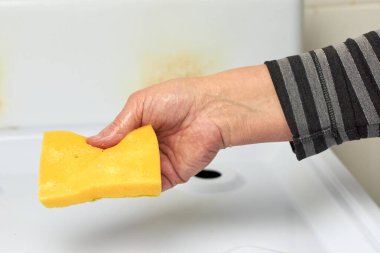 Bir kadının eli çizgili kolunda, gaz sobasını yıkamak için ıslak, sarı bir bulaşık süngeri tutar.