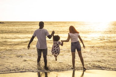 Çok ırklı bir aile sahilde eğleniyor. Baba, anne ve kız sahilde birlikte oynuyorlar. Tatil ve seyahat kavramı.