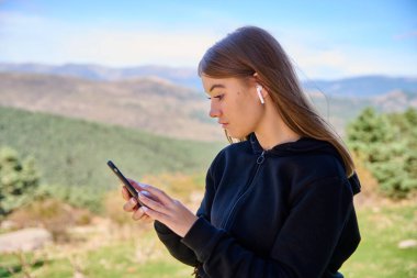 Kafkasyalı bir kadın müzik dinliyor ve arka planda yeşil tepeler olan telefonunda daktilo yazıyor.
