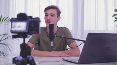 Blogcu adam video bloğunu evde kaydediyor. Genç çocuk video blogunu kaydediyor. Genç adam blog kanalı için bir video kameranın önünde konuşuyor. Vlog kaydedicisi akıllı telefon kullanarak çevrimiçi yayın yapıyor 