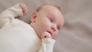 Güzel bebek. Mavi elbiseli sevimli iki aylık bebek kameraya bakıyor, ağır çekimde. Aktif yeni doğan çocuk kanepeye uzanıyor ve kollarını hareket ettiriyor. Tombul bir erkek bebeğin yüksek açısı.