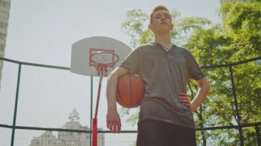 Basketbol sahasında duran ve elinde basketbol topu tutan bir çocuk. Elinde top, arkasında basketbol potası olan bir gencin sinematik çekimi. Kendine güvenen bir basketbolcunun videosu. Kol altında basketbol.