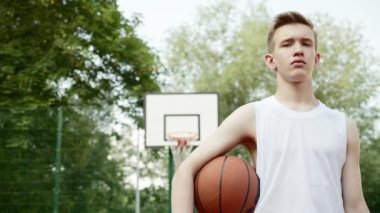 Basketbol sahasında duran ve elinde basketbol topu tutan bir çocuk. Elinde top, arkasında basketbol potası olan bir gencin sinematik çekimi. Kendine güvenen bir basketbolcunun videosu.. 