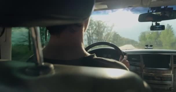 若い男へのリアビューは車を運転します 10代の若者は日没時に車を運転する 若い男が車を運転している 十代の少年は車を運転する 太陽の光は車のフロントガラスを通して輝きます リアルタイム — ストック動画
