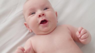 Yeni doğmuş güzel bir bebek. Çıplak göbekli, iki aylık tatlı bir bebek kameraya bakıyor, ağır çekimde. Aktif yeni doğan çocuk yatağa uzanıyor ve kollarını hareket ettiriyor. Tombul bir bebeğin yüksek açılı görüntüsü.