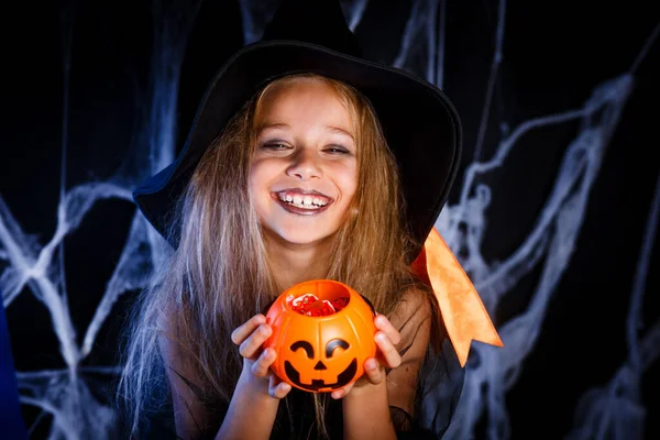 Nettes glückliches kleines Mädchen verkleidet als Hexe mit einem Eimer Süßigkeiten auf Halloween-Hintergrund — Stockfoto