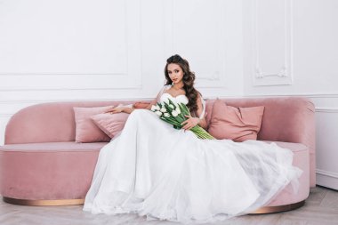 Gelin, elinde bir buket düğün elbisesi ve içeride bir kanepede oturuyor. Düğün günü.