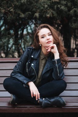 Siyah ceketli genç, kıvırcık bir kadın açık havada bir bankta oturuyor.