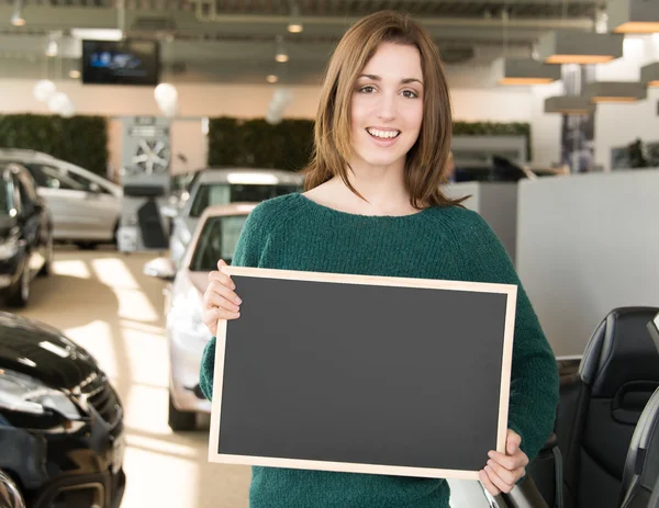 Jeune brune tenant tableau noir à l'intérieur concessionnaire automobile — Photo