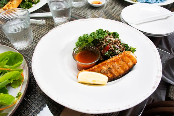 Thai chef cooking food fusion style cuisine roasted grilled fried Salmon steak healthy with spicy sauce and vegetables fruits salad on ceramic dish plate served at restaurant cafe in Bangkok, Thailand