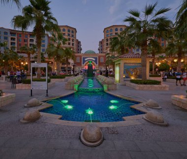 Peral Doha 'daki Medina Merkezi, Katar' ın günbatımı, arka planda Souq Almedina 'yı ön planda aydınlatılmış bir çeşmeyle, palmiye ağaçları ve yürüyen insanlarla gösteriyor.