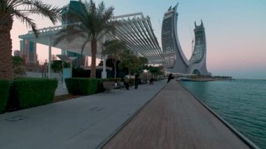 Lusail Corniche, Lusail City 'deki marinada, Katar' da gün batımı çekimleri fıskiyeyi gösteriyor, insanlar arka planda ufuk çizgisiyle geziniyor.