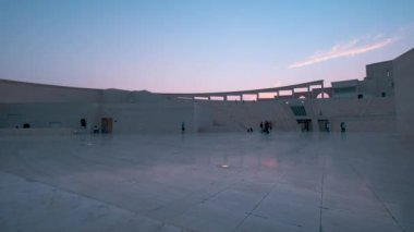 Doha 'daki Katara Kültür Köyü, Katar' ın gün batımında, arka planda yerli halk ve ziyaretçilerin yürüdüğü, oturduğu ve bulutların olduğu amfitiyatronun çekimini gösteriyor.