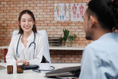 Beyaz tişörtlü, Asyalı bir doktor olan güzel bir kadın doktor, duvarın arkasındaki klinikteki erkek hastayı muayene ediyor, tıbbi uzmanlık alanında gülümsüyor..