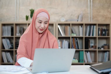 Asya kökenli güzel bir iş kadını, küçük işletme ofisinde dizüstü bilgisayar ve internet iletişimi kullanarak e-ticaret üzerinde çalışıyor. Çekici kişi, geleneksel İslam tesettüre göre giyinir..