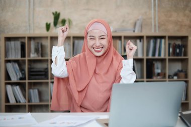 Masasında dizüstü bilgisayarı olan güzel bir İslamcı kadın kurucusu küçük ofis e-ticaret şirketlerinin başarısını kutlamak için ellerini kaldırıyor. Genç müdür gülümsüyor ve mutlu çalışıyor.
