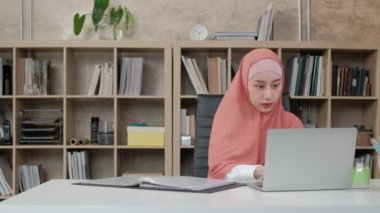 Asya kökenli güzel bir iş kadını, küçük işletme ofisinde dizüstü bilgisayar ve internet iletişimi kullanarak e-ticaret üzerinde çalışıyor. Çekici kişi, geleneksel İslam tesettüre göre giyinir..