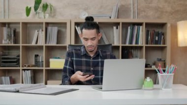 Asya kökenli genç bir erkek işçi, yaratıcı işler yapmak için dizüstü bilgisayar, masa başında cep telefonu iletişimi, günlük iş yerinin arkasında kitaplık, girişim işçisi ve çevrimiçi e-ticaret mesleği.