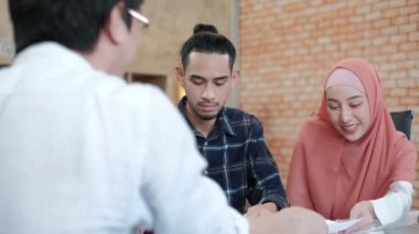 İslami kökenli iki genç iş arkadaşı, gülümseyen bir müşteriyle finansal projeler hakkında konuşuyor, küçük bir ofis işyerindeki çalışma masasında bir iş grafiğiyle başarı sunuyor..