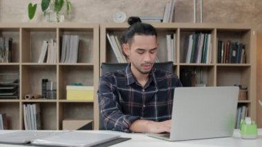 Asya kökenli genç erkek işçi, günlük bir iş yerinin, girişim işadamının ve çevrimiçi e-ticaret mesleğinin kitaplığının önündeki beyaz masada yaratıcı işler yapmak için dizüstü bilgisayar kullanıyor..
