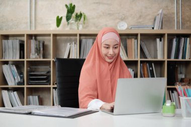 Asya kökenli güzel bir iş kadını, küçük işletme ofisinde dizüstü bilgisayar ve internet iletişimi kullanarak e-ticaret üzerinde çalışıyor. Çekici kişi, geleneksel İslam tesettüre göre giyinir..
