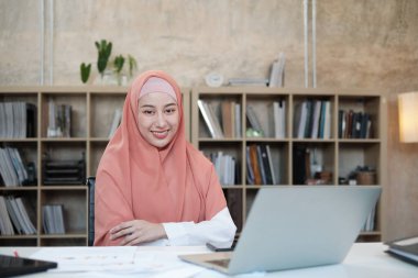 İslami kadın tesettürlü, tesettürlü ve küçük bir ofis iş yerinde kameraya bakan, beyaz masadaki dizüstü bilgisayarla çalışan, arkasında kitaplık olan güzel bir iş adamının portresi..
