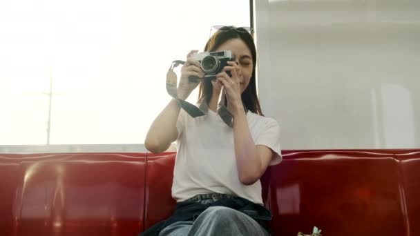 美しいアジアの女性観光客とカメラは赤い座席に座って 空の列車で移動し 都市の景色 鉄道で都市の乗客のライフスタイル 幸せな旅の休暇で輸送するときにスナップショット写真を撮る — ストック動画