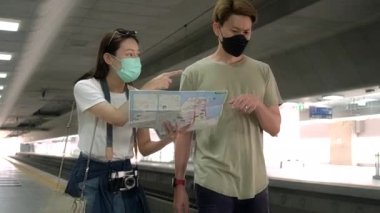 Yüz maskeli genç bir çift bilgi arıyor, Tayland 'daki tren istasyonunda seyahat için konum, yolcu yolculuğu yaşam tarzı, seyahat tatilinde günlük ulaşım. 