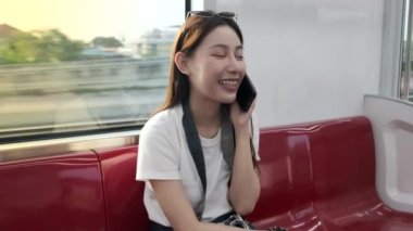 Kameralı güzel Asyalı bayan turist kırmızı koltukta oturur, gökyüzü treniyle seyahat eder, şehir manzarasında taşınırken cep telefonuyla konuşur, tren yoluyla şehir yolcusu yaşam tarzı, iyi yolculuklar..