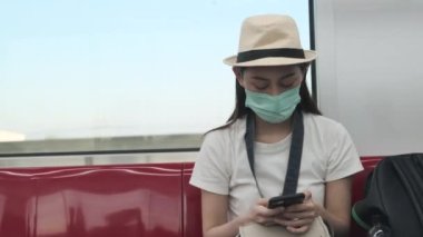 Yüz maskeli, şapkalı Asyalı kadın turist gökyüzü treninde oturuyor, cep telefonuyla mesajlaşıyor, demiryolu ile seyahat ediyor, şehir yolcusu yaşam tarzı, Tayland 'da yeni normal bir seyahat tatili..