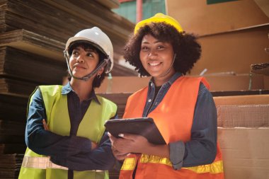 Güvenlik üniforması ve kask stoku kontrolü yapan iki kadın işçi ve iş arkadaşı bir sürü kağıt yığını olan depo fabrikasının deposunda kontrollü üretim yapıyorlar. Arkadaşı geri dönüşüm imalat işinde çalışıyor..