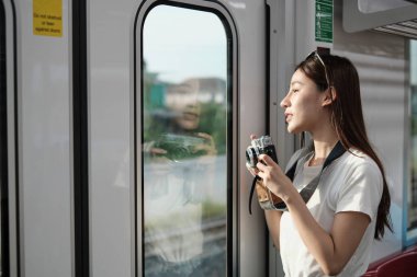 Yolcu kabininde kameralı güzel bir Asyalı kadın turist, gökyüzü treniyle seyahat ediyor, şehir manzarasında taşınırken fotoğraf çekiyor, demiryolu ile şehir hayatı, iyi yolculuklar..