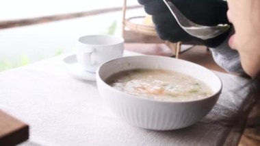 Lezzetli kış geleneksel Tayland kahvaltısı yemeği. Asyalı bir kadının, eldivenli, beyaz bardak sıcak pirinç, domuz eti ve sisli bir sabah kaşığıyla haşlanmış yumurtayı yerken, sağlıklı ve lezzetli yiyecekler yerken yakın plan fotoğrafı..