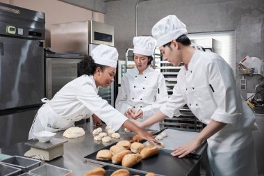 Profesyonel gurme takımı, beyaz aşçı üniforması ve önlüklü üç genç aşçı hamur ve yumurta yoğurur, ekmek ve taze fırın yemeği pişirir paslanmaz çelik restoran mutfağında fırında pişirir..