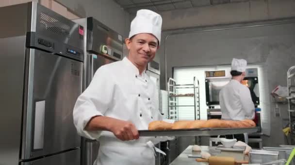 身穿白衣 头戴礼帽 面带微笑地展示新鲜美味面包的资深亚洲男性厨师 看着相机 对自己的烘焙食品感到满意 并在不锈钢厨房担任专业工作 — 图库视频影像
