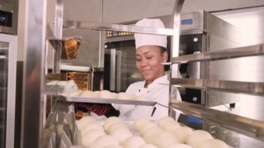 Profesyonel Afro-Amerikan kadın aşçı üniforması, eldiven ve önlük, hamur işiyle ekmek yapmak, taze fırın yemekleri hazırlamak, restoranın paslanmaz çelik mutfağında fırında pişirmek..